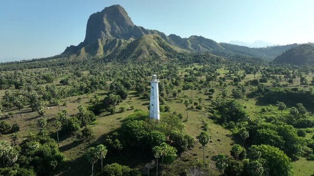 Aerial view of the white Nuca Molas lighthouse surrounded by palm trees and grassy fields with a mountain backdrop in Satar Mese Barat, East Nusa Tenggara, Indonesia.