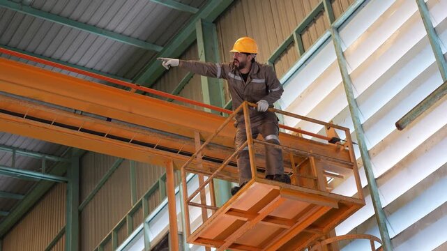 Industrial worker standing on crane platform pointing forward in factory, Factory operator inspecting overhead crane in manufacturing plant, Technician standing on maintenance platform in facility