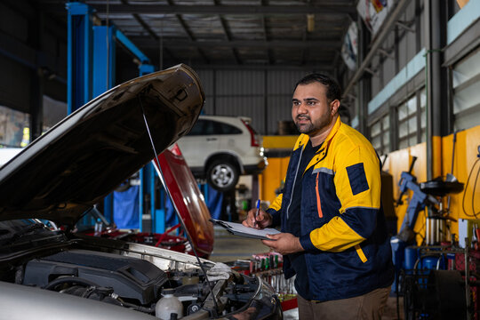 asian mechanic in workwear writing paper on clipboard after inspection of a car engine that needs to be repaired in order to provide a quotation to customer