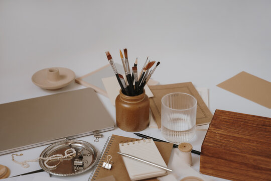 Neutral Warm Workspace Arrangement Featuring Kraft Paper Journals, Silver Laptop, Glass Of Water, And Studio Accessories