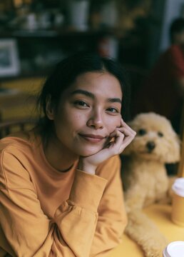 Portrait of a young woman sitting at a table with a small dog. the woman is wearing a yellow long-sleeved shirt and has shoulder-length dark hair.