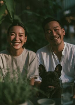 Young couple sitting at a table with a black french bulldog in front of them. they are both smiling and looking at the camera.