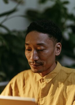 Close-up portrait of a middle-aged asian man. he is wearing a yellow collared shirt and is sitting in front of a laptop.