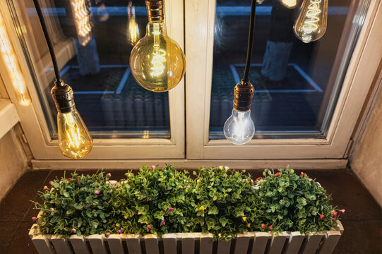 Hanging light bulbs shine above green plants in a windowbox. The scene shows a balcony with bright lights illuminating the night