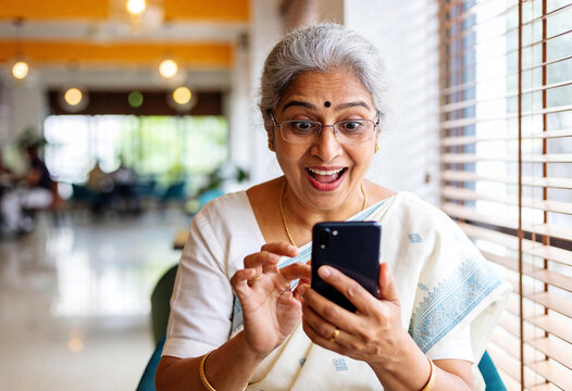 Senior Indian woman surprised looking at smartphone in bright modern cafe wearing traditional white saree and bindi