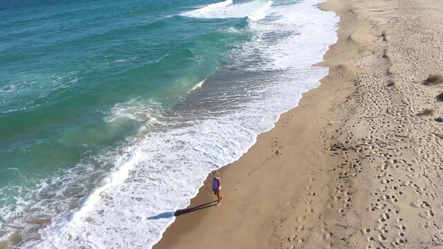 Slow motion aerial view of a gorgeous blonde woman walking on a beach. Sea waves washing sandy shoreline