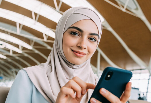 Smiling young Muslim woman wearing beige hijab using smartphone in modern airport terminal with natural lighting