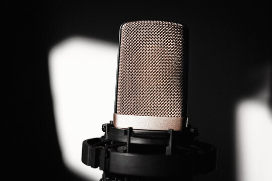 Studio microphone with large diamond mesh pop filter grille, lit against black background with soft light patches behind. Shock mount cradles its base giving setup a professional recording studio look