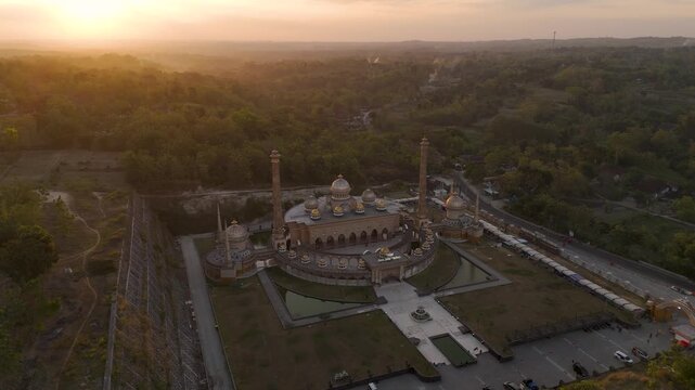 Aerial view of Masjid Samin Baitul Muttaqin featuring golden domes and minarets surrounded by lush greenery during a warm sunset in Bojonegoro, East Java, Indonesia.