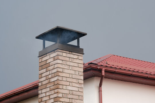 Smoke rising from a brick chimney on a house with a red roof against a gray sky