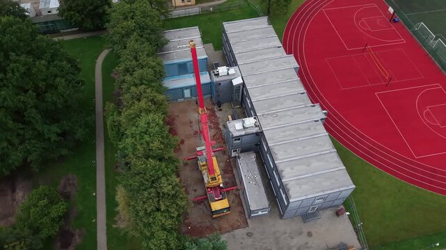 Aerial footage captures a construction crane assembling modular buildings beside a school sports field and running track, surrounded by lush green trees on a cloudy day.