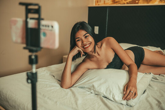 Smiling adult woman in lingerie poses on a bed for a smartphone camera, illustrating subscription-based creator content, online modeling, fan engagement, digital income, and sensual lifestyle media.
