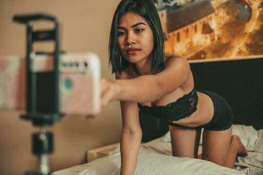 Adult woman in lingerie adjusts a smartphone mounted beside her bed before recording premium subscriber adult video content.