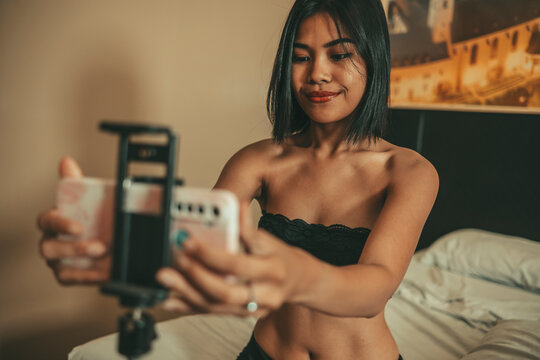 Adult woman wearing lingerie operates a smartphone on a bedside mount while preparing to film subscriber content, representing premium digital media and independent content production.