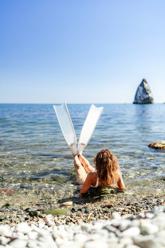 White swim fins extend upward while a woman rests in clear shallow water near a rocky coastal landscape