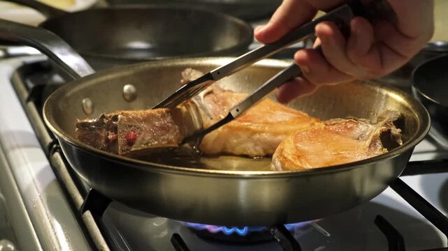 Flipping of roast pork steak in a steel pan in kitchen stove, cooking with metal frying pan, stove surface texture, from a close-up perspective, roasting until golden brown. Rotating with a spatula.