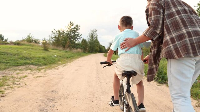 Mother supports son as he wobbles on bicycle along sandy village road. He repeatedly loses balance yet keeps learning with her steady encouragement and care.