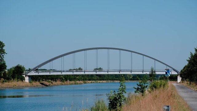 A beautiful architectural view of a tied-arch bridge crossing a peaceful shipping canal under a clear blue sky.