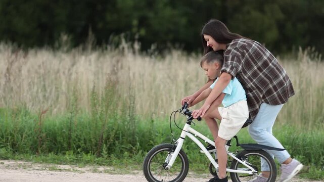 Mother and son ride white bicycle along sandy road in rural countryside. Side view follows their movement while open field passes softly in background.
