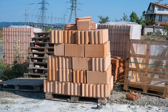 A stack of terracotta hollow clay bricks sits on a wooden pallet at an outdoor construction site on a clear, sunny day.