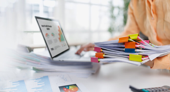 Businesswoman in a mustard sweater organizes colorful paperwork on her desk, using a laptop, showcasing a busy and productive work environment with office supplies.