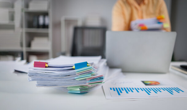 Businesswoman in a mustard sweater organizes colorful paperwork on her desk, using a laptop, showcasing a busy and productive work environment with office supplies.