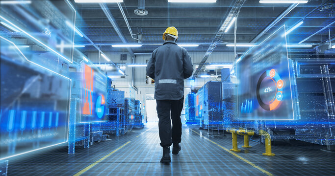 Rear View of a Male Factory Engineer Walking Down a Modern Plant Aisle Surrounded by Holographic Analytics Dashboards, Ar Charts and Digitized Industrial Machines Visualizing Production Monitoring.