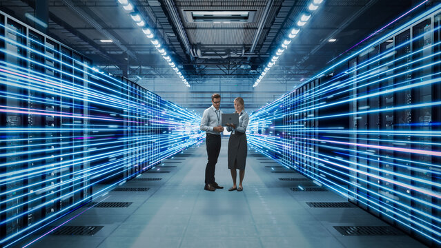 Two Caucasian It Professionals, a Man and a Blonde Woman, Review Network Data on a Laptop While Standing Between Server Racks Emitting Fast Neon Blue Fiber Optic Light Trails.