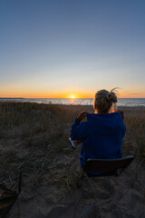 Fototapeta premium Rear view of young blonde woman in blue hoodie sitting on camping chair relaxing on sandy beach watching sunset over Baltic sea in Nova Estonia.