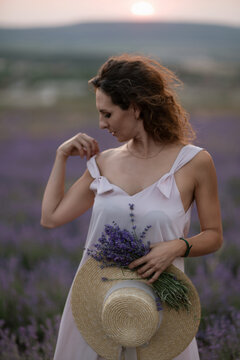 Summer dress woman holding lavender bouquet and straw hat in purple flowering Lavandula field during sunset