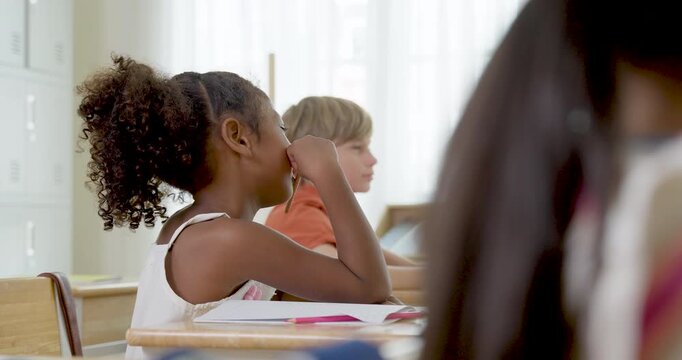 Two young girl student sit at wooden desks with notebooks, listening to teacher while joining primary classroom discussion, peer interaction, classwork routine and back to school learning moment