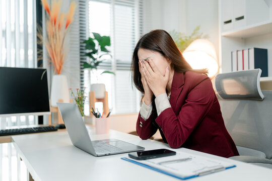 Stressed young asian businesswoman feeling overwhelmed and frustrated, suffering from burnout while working in a modern office, covering her face in despair
