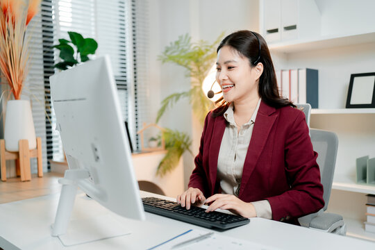 Professional asian woman working at call center or customer support office, wearing a headset, smiling and typing on computer keyboard, providing online assistance and communication