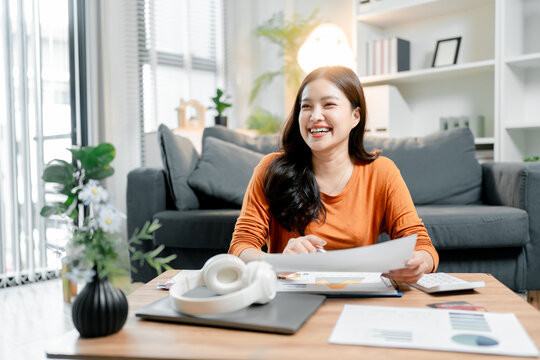 Smiling young asian woman holding financial documents, managing personal finances or remote business operations from her home office, enjoying work with a calculator and laptop on the table
