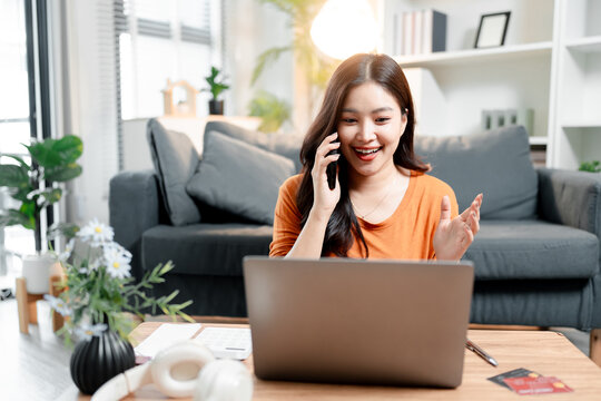 Smiling young asian woman having a cheerful conversation on her smartphone while working on a laptop at home, discussing business, shopping online, and managing finances