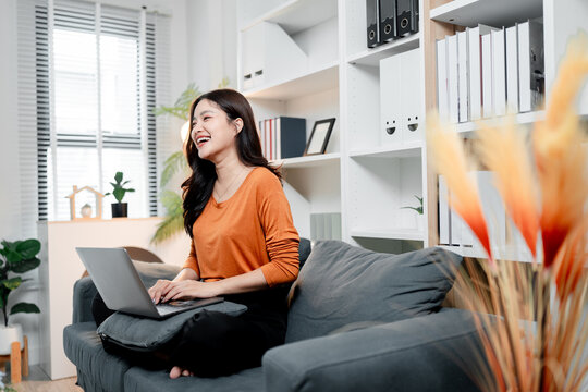 Young asian woman laughing and working remotely on laptop in a comfortable living room, enjoying her freelance lifestyle while achieving productivity and happiness at home