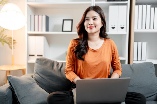 Young asian woman sitting on sofa at home, using laptop for remote work or online learning, smiling while thinking about future business and freelance opportunities