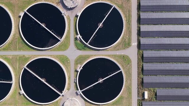 Aerial view of a wastewater treatment plant featuring four large circular sedimentation tanks and adjacent rows of solar panel arrays in Den Hoorn, South Holland, Netherlands.