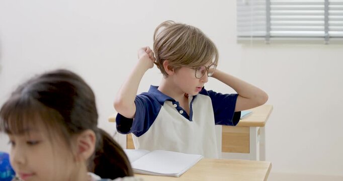 Elementary boy with glasses thinks, adjusts hair, raises pencil during classroom question time while asian girl studies nearby, back to school activity builds confidence, answer readiness, attention