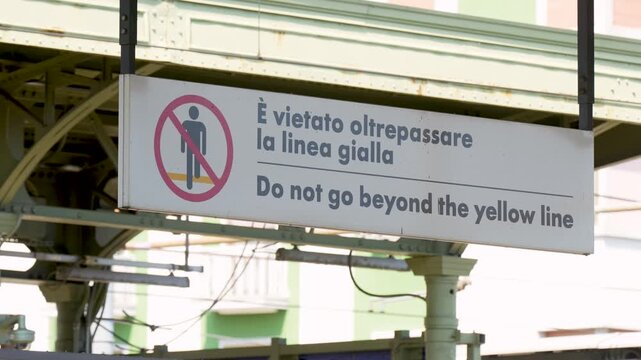 Sign prohibiting crossing the yellow line at a train station, featuring bilingual instructions in Italian and English, with a clear view of the surrounding urban environment