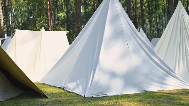 Various white tents are arranged on green grass in a wooded area. The setting shows bright sunlight filtering through the trees, creating patterns on the ground
