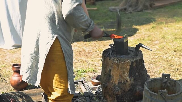 A person is working at a forge outdoors, using heat and tools to shape metal. The setting features natural surroundings and rustic materials, showcasing traditional craftsmanship
