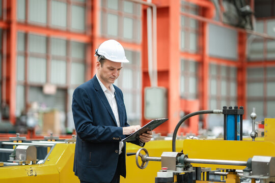 Industrial manager checking production data on clipboard beside factory machinery, representing manufacturing management, engineering inspection, quality assurance, productivity.