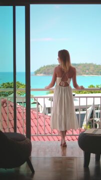 Young woman in white dress stand on balcony enjoy ocean view. Female relax in Phuket, Thailand exotic nature. Outdoor lifestyle travel summer holiday vacation on tropical island. Back view slow motion