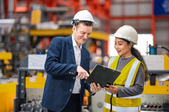 Factory manager points at clipboard while discussing production information with female engineer, industrial teamwork, manufacturing management, quality control.