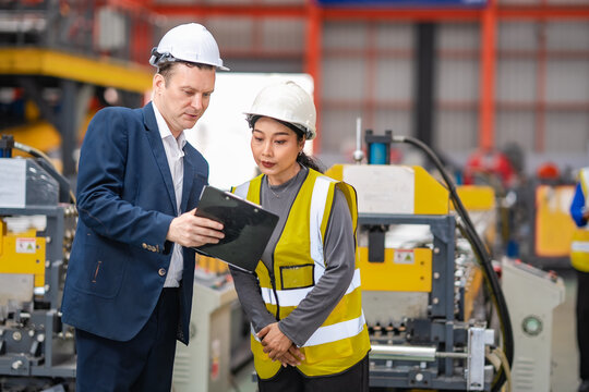 Factory manager and female engineer review clipboard together beside production machinery, discussing manufacturing operations, inspection results, quality control.
