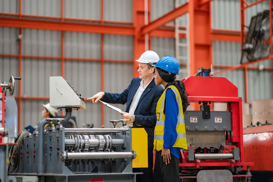 Factory manager explains machine control system to female engineer on production floor, industrial equipment operation, manufacturing process, workplace training.