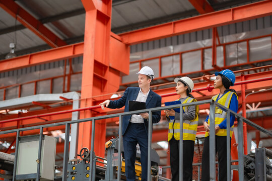 Factory manager leads two female engineers during factory inspection, discussing production operations from elevated platform, industrial teamwork, manufacturing management.