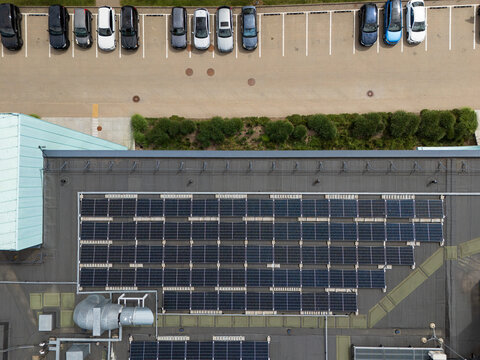 Aerial top-down view of solar panels installed on commercial building rooftop beside parking lot with cars, urban renewable energy infrastructure concept.
