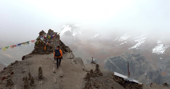 Annapurna Circuit, Manang region, Nepal, orbit drone shot of a male trekker standing at High Camp View Point beside stone cairns and prayer flags on the route to Thorong La.